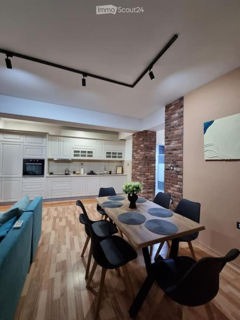 Modern open-plan living and dining area with a wooden dining table, black chairs, a blue sofa, white kitchen cabinets, and a brick accent wall.