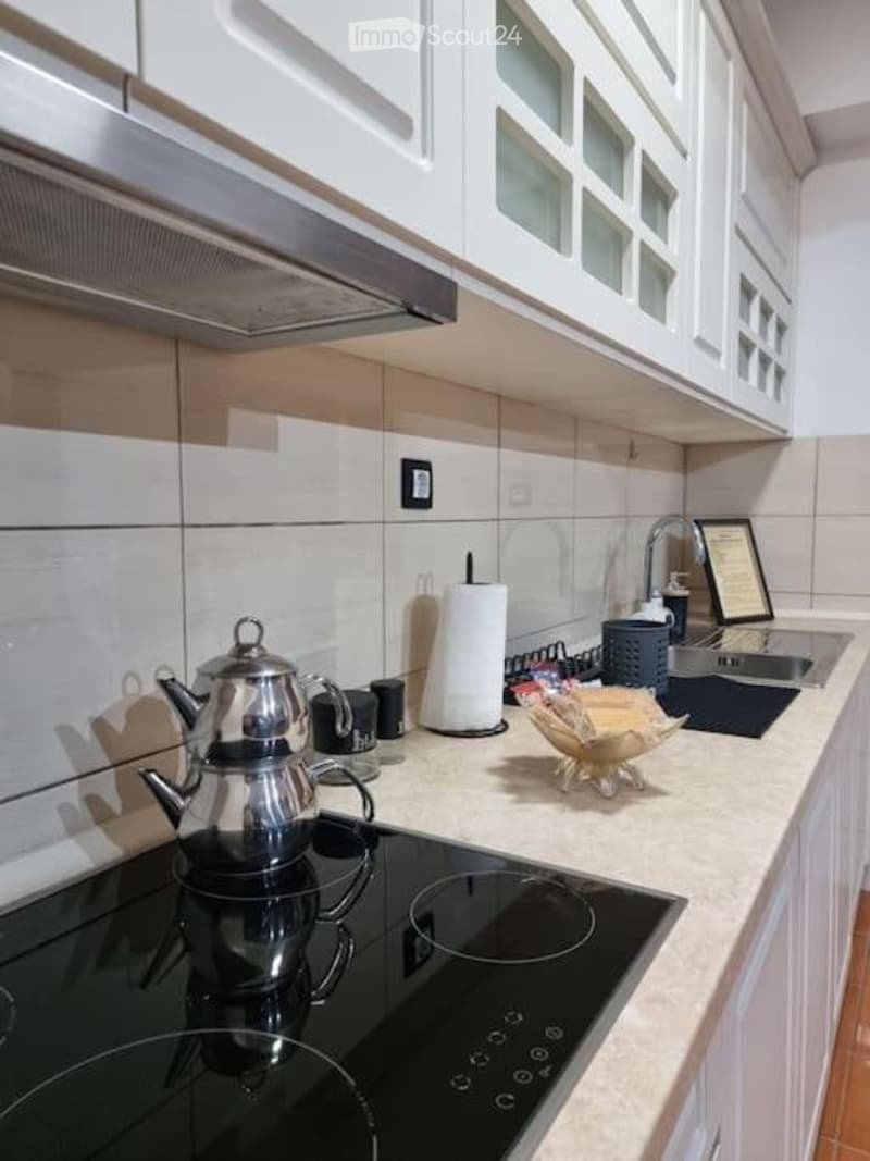 Modern kitchen with white cabinets, stainless steel appliances, and a black electric stove. Includes a teapot, paper towel, and a decorative bowl on the counter.