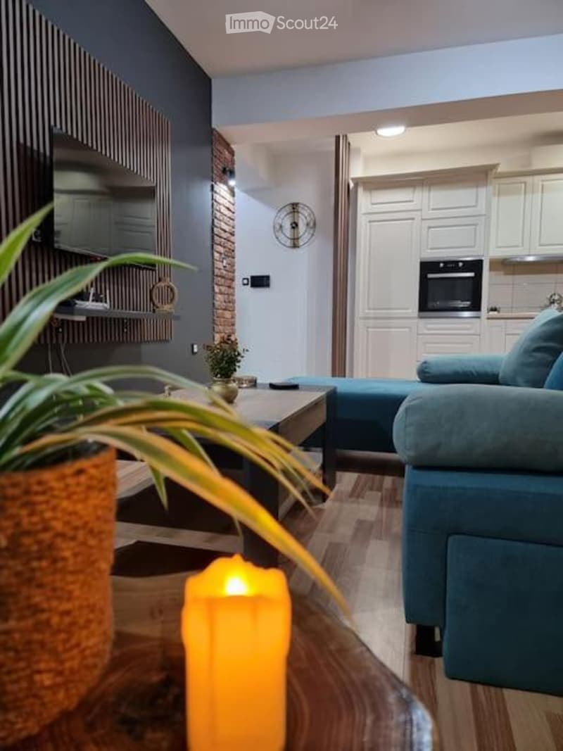 A modern living room with a blue sofa, coffee table, and a small table with a candle. The room features a brick accent wall, white cabinets, and a small plant on a shelf.