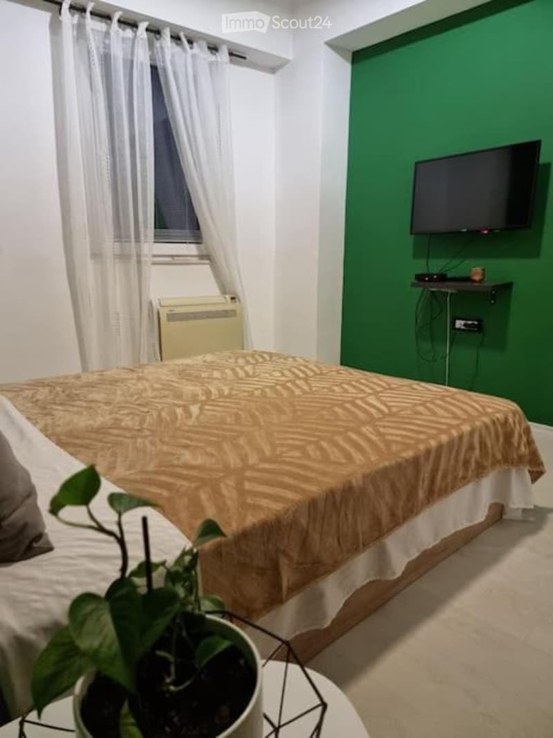 Cozy bedroom with a double bed, patterned bedspread, a potted plant on a small table, a wall-mounted TV, and a window with white curtains.
