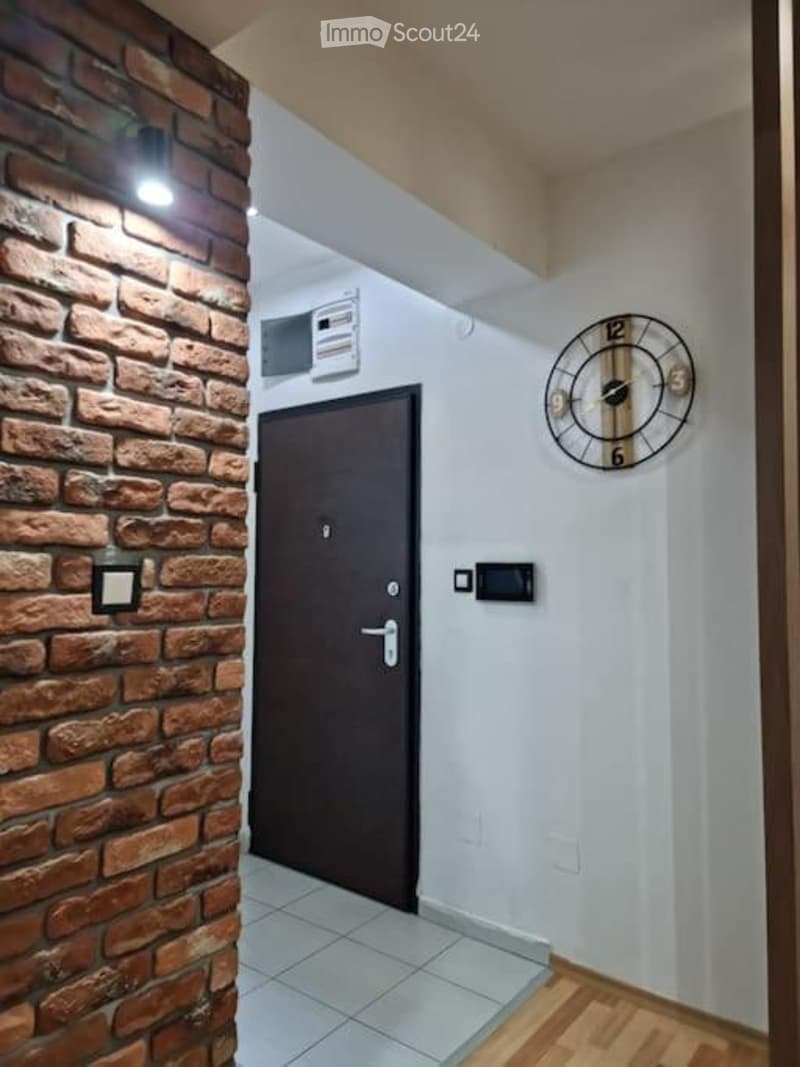 A narrow hallway with a brick accent wall on one side and a wooden floor on the other. Features include a closed door, a wall-mounted clock, and several light switches.