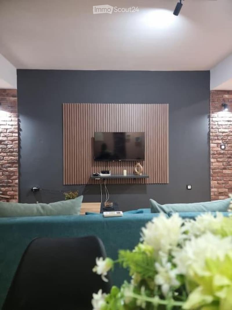 Modern living room with a dark blue accent wall, a teal sofa, a mounted TV, and a decorative wall-mounted shelf. The space features a brick accent wall and a sleek, contemporary design.