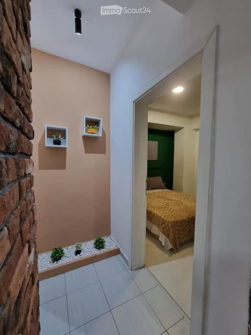 A modern hallway with a brick accent wall, white floating shelves with small plants, and a view into a furnished bedroom with a bed and patterned bedding.