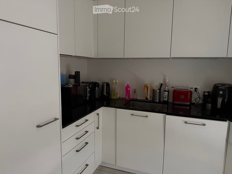 Modern kitchen with white cabinets, black countertops, and various appliances including a fridge, microwave, toaster, and coffee maker.