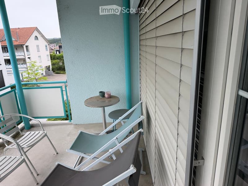 Balcony with a small round table, three chairs, and a view of neighboring buildings and greenery.