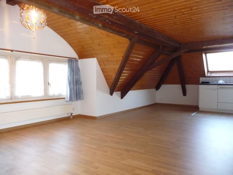 Empty attic space with wooden beams, wooden flooring, white walls, and a small kitchen area with white cabinets and a sink.