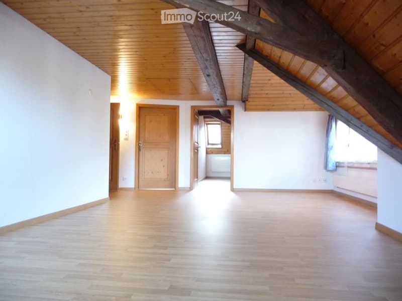 Empty attic room with wooden floors, exposed wooden beams, and white walls. Features a wooden door and a small window with a curtain.