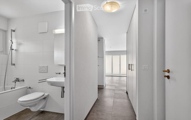 Modern bathroom with white tiles, bathtub, toilet, sink, and shower. The hallway leads to another room with large windows.