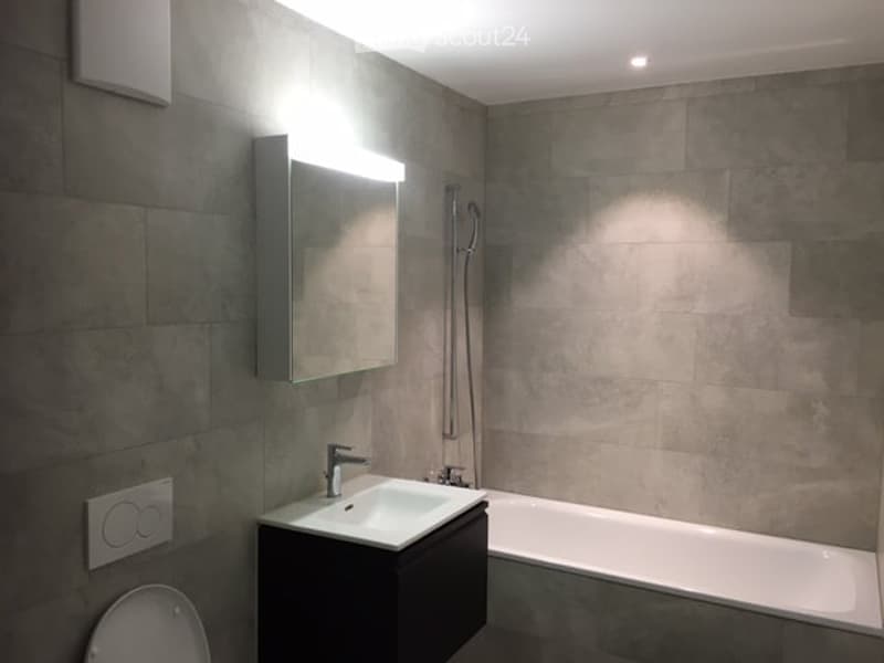 Modern bathroom with grey tiled walls, a black vanity with a white sink, a large mirror with integrated lighting, and a white bathtub.