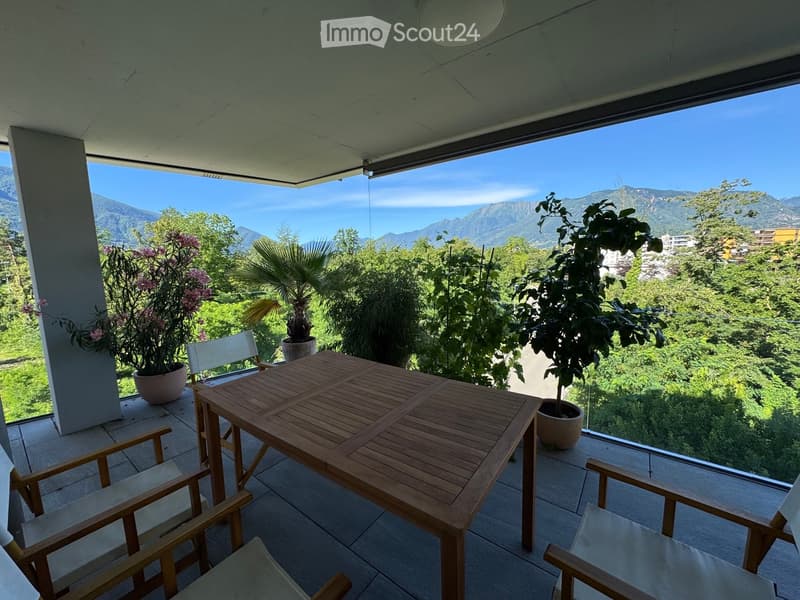 Balcony with wooden table and chairs, potted plants, and scenic mountain view.