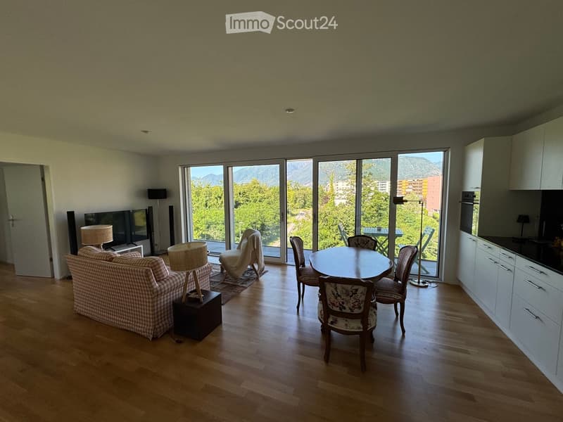 Bright living and dining area with wooden flooring, large windows offering a scenic mountain view, a sofa, a dining table with chairs, and lamps.