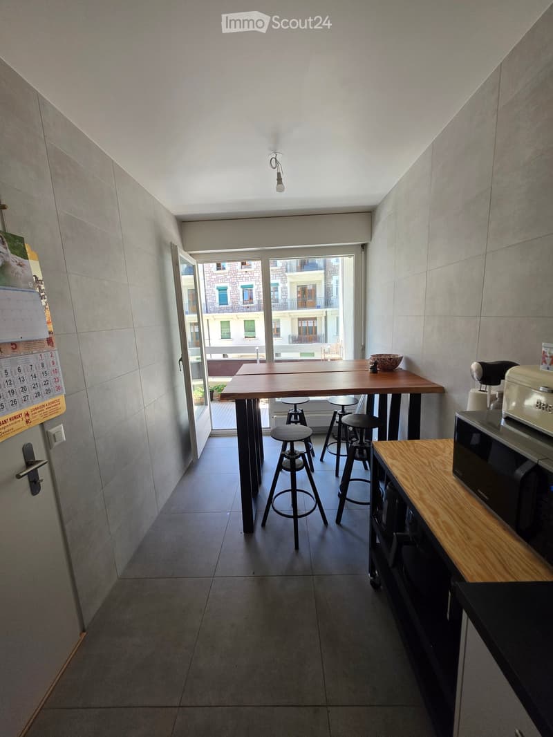 Modern kitchen with a large wooden dining table, four stools, microwave, and a bowl on the table. The room has large windows with a view of the city and a calendar on the wall.