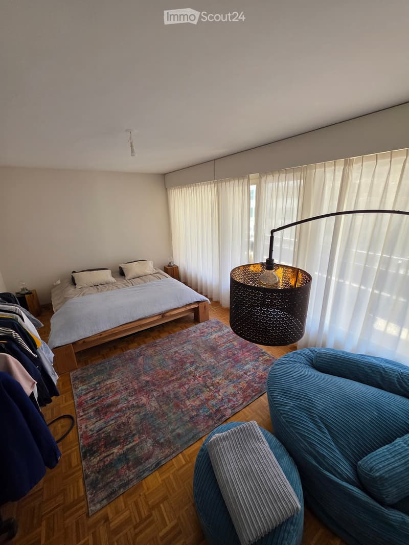 A modern bedroom with a wooden bed, a blue velvet sofa, a colorful rug, and a floor lamp with a decorative shade. The room features large windows with white curtains and wooden flooring.