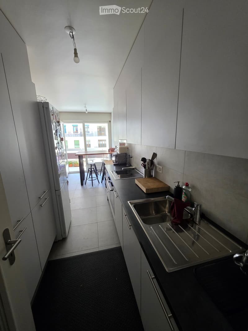 Modern kitchen with white cabinets, stainless steel sink, and black countertops. Includes a refrigerator, cutting board, and a small dining area with stools visible through an open doorway.