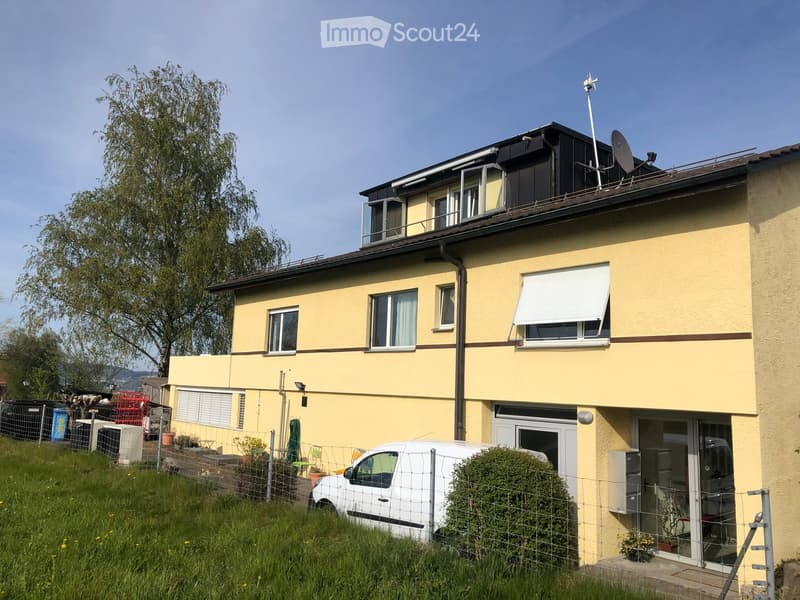 Two-story yellow house with multiple windows, a raised ground floor, and a satellite dish on the roof. The house features a balcony on the upper level and a fenced garden area with a parked van.