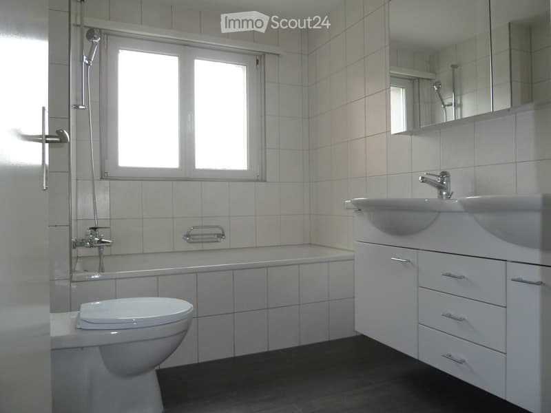 Modern bathroom with white tiles, a toilet, a sink with a mirror, a bathtub, and a window for natural light.