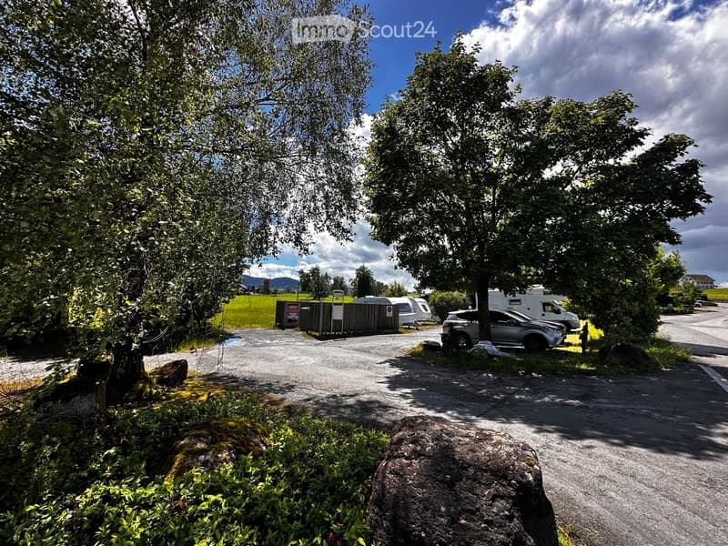 A gravel parking area surrounded by trees and greenery, with several parked vehicles including a camper van and a car, and a fenced area in the background.