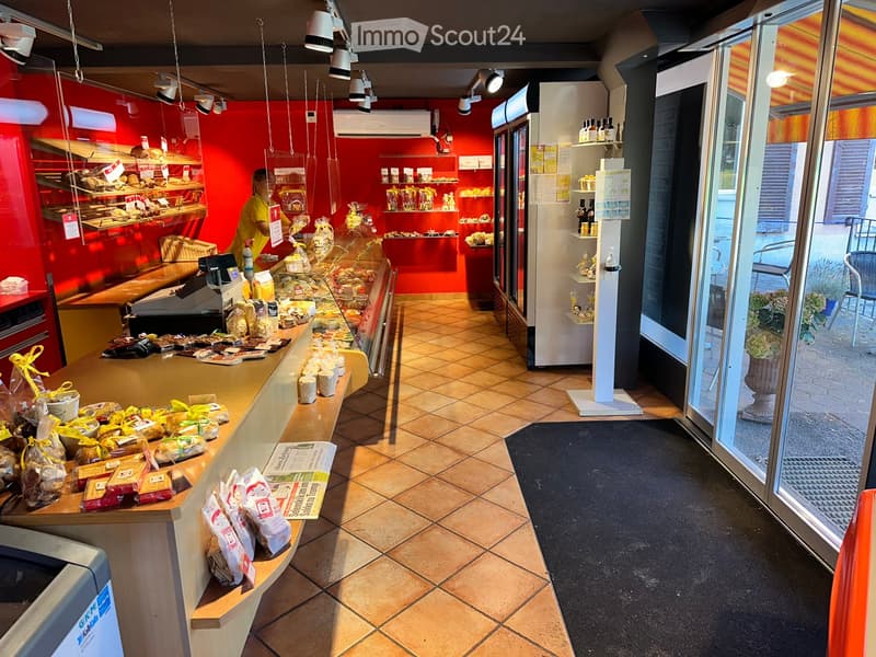 Bakery interior with red walls, tiled floor, counter displaying bread and pastries, shelves with products, and a refrigerator.