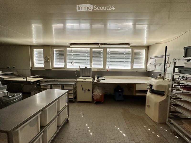 A clean, well-lit kitchen with stainless steel counters, cabinets, and various appliances including an oven and refrigerator. The space features multiple windows with blinds and a tiled floor.
