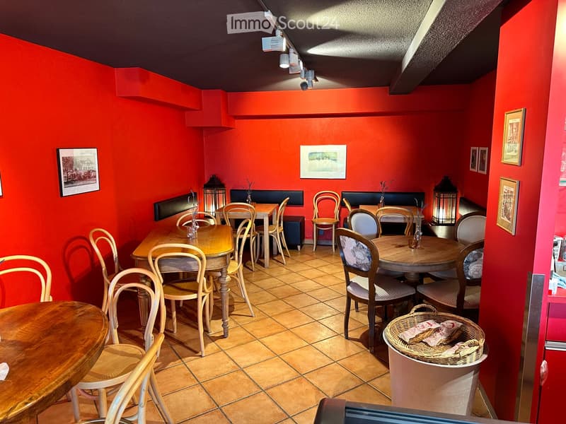A cozy restaurant with red walls, wooden tables, and chairs arranged around the space. Lanterns and picture frames adorn the walls, and a basket with menus sits on a counter.
