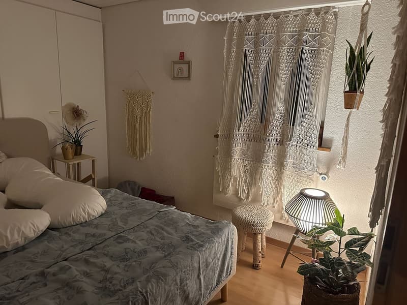 Cozy bedroom with a bed, nightstand, hanging plants, and decorative wall hangings.