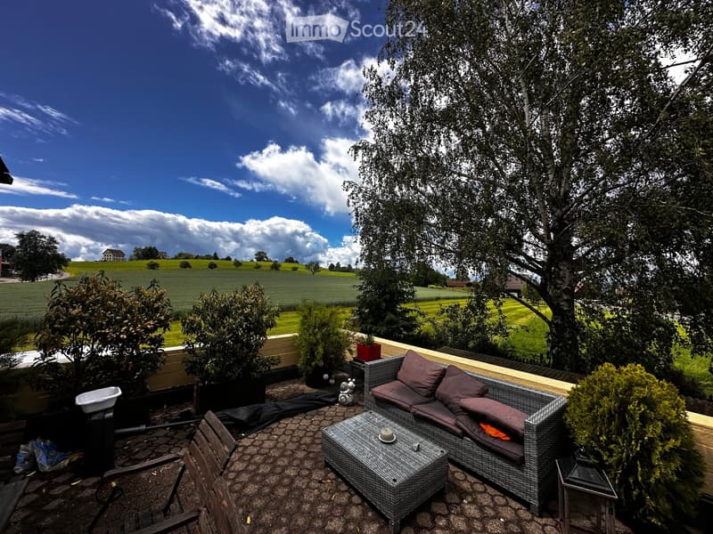Balcony with wicker sofa, coffee table, and wooden chairs, surrounded by greenery and a scenic countryside view.