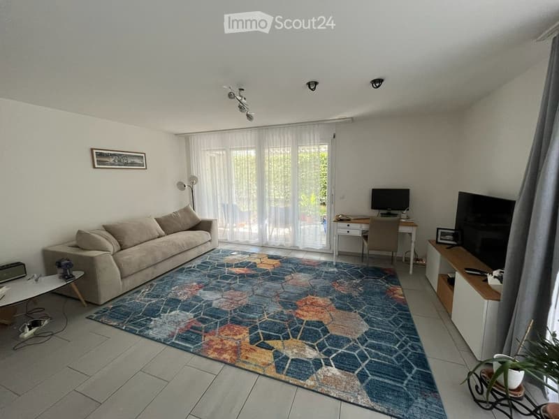 Bright living room with a beige sofa, colorful geometric rug, coffee table, desk with chair, television, and floor lamp. Large windows with sheer curtains let in natural light.