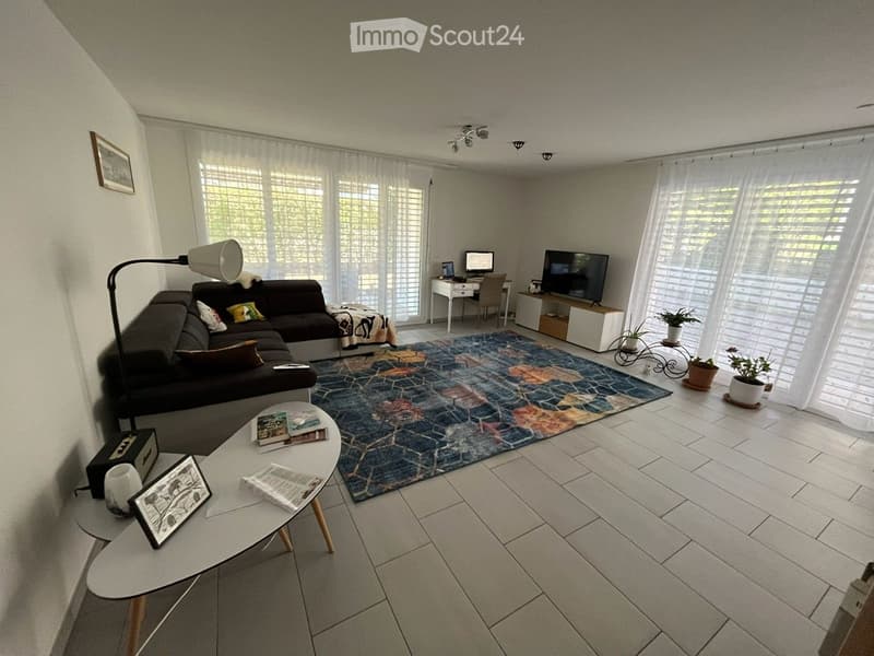 Modern living room with white tiled flooring, large windows with blinds, a dark grey sectional sofa, a colorful patterned rug, a white coffee table with magazines, a floor lamp, a TV stand with a flat-screen TV, a small desk with a laptop, and potted plan