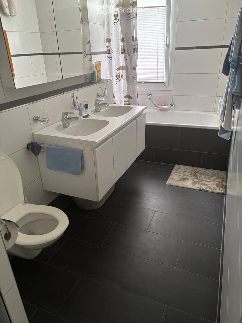 Modern bathroom with black tiled floor, white sink with dual faucets, toilet, bathtub, mirror, and towels.