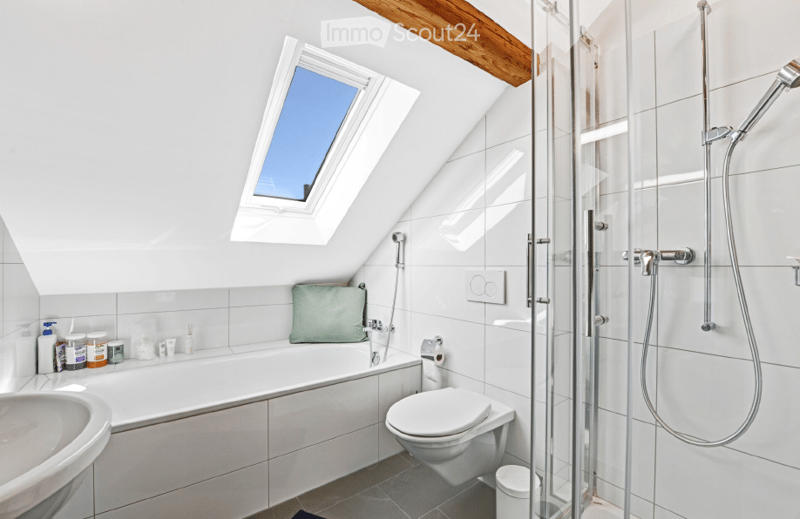 Modern bathroom with white tiles, a bathtub, toilet, sink, and a glass-enclosed shower. Features a skylight window and a small decorative pillow on the bathtub.