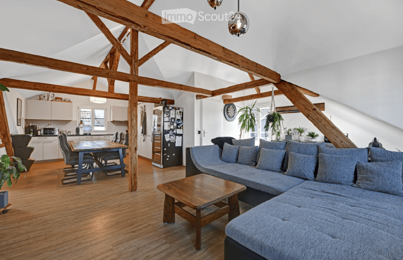 Open-plan living and dining area with exposed wooden beams, grey sectional sofa, wooden coffee table, dining table with chairs, kitchen area with cabinets and appliances.