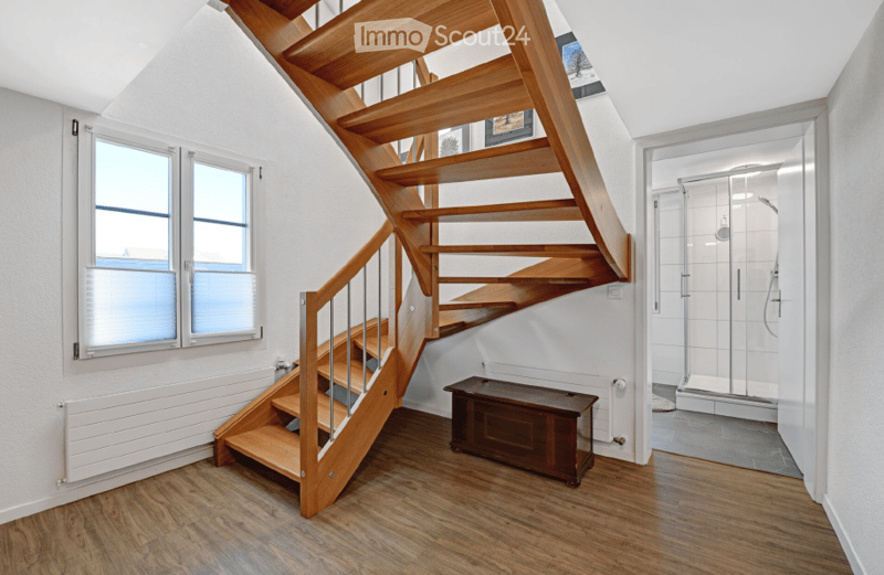 A modern hallway with wooden flooring, a wooden staircase with a metal railing, a small wooden bench, and a glass-enclosed shower area visible in the background.