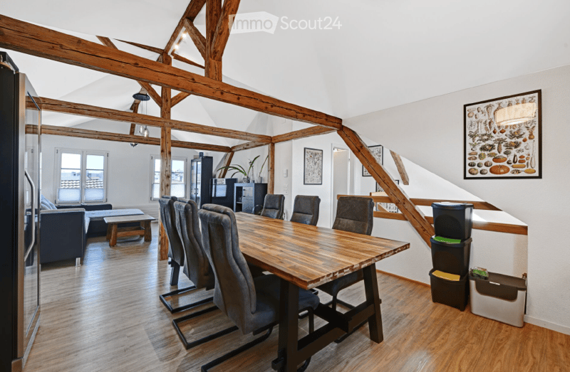 Open-plan living and dining area with exposed wooden beams, hardwood flooring, a large wooden dining table with six chairs, a sofa, coffee table, and a refrigerator. The space is well-lit with large windows and features decorative artwork on the walls.