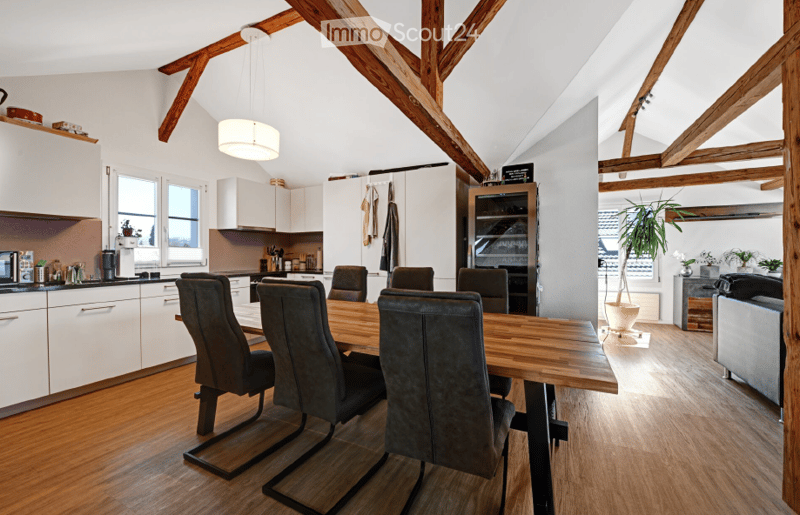 Open-plan kitchen and dining area with exposed wooden beams, modern white cabinets, a wooden dining table with six chairs, and a small living area with a sofa and potted plant.