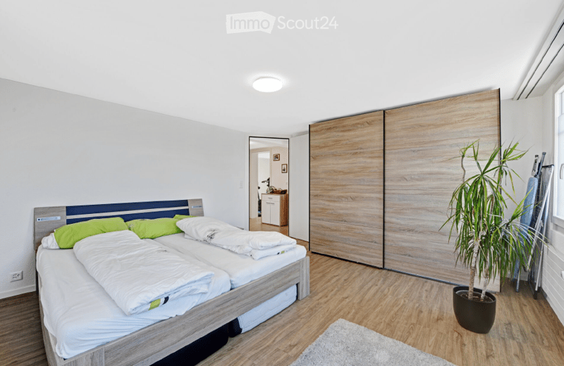 Modern bedroom with a double bed, green pillows, a large wooden wardrobe, and a potted plant in the corner.