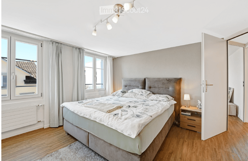 Modern bedroom with a large bed, nightstand, lamp, and curtains. The room features wooden flooring and large windows with a view of the roof.