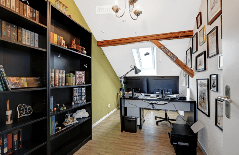 A modern, well-lit room with wooden floors, a large bookshelf filled with books and decorative items, a sleek white desk with a laptop and office chair, and several framed pictures on the wall. The room features exposed wooden beams and a skylight, creati