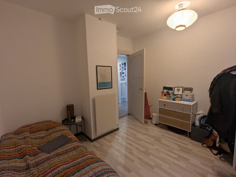 Cozy bedroom with wooden flooring, a bed with a colorful striped blanket, a nightstand, a small table with a chair, a radiator, a cabinet with drawers, and a framed picture on the wall. An open door leads to another room.