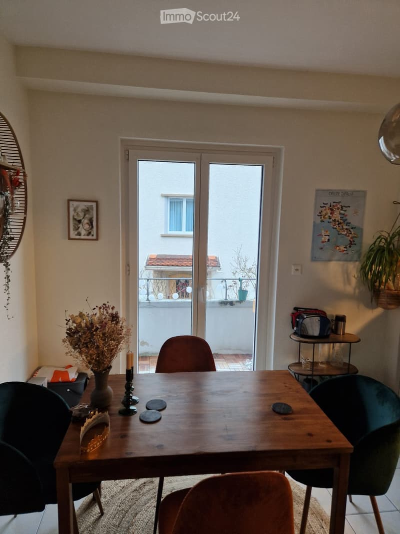 Dining area with wooden table, chairs, vase with dried flowers, candles, and a small table with a toaster. A sliding glass door leads to a balcony with a view of the building.