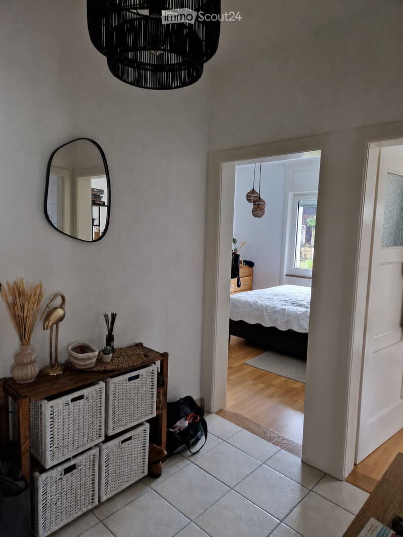 Hallway with a large black hanging light fixture, a round mirror on the wall, and a wooden shelf with decorative items. An open doorway leads to a bedroom with a bed, wooden flooring, and hanging lights.
