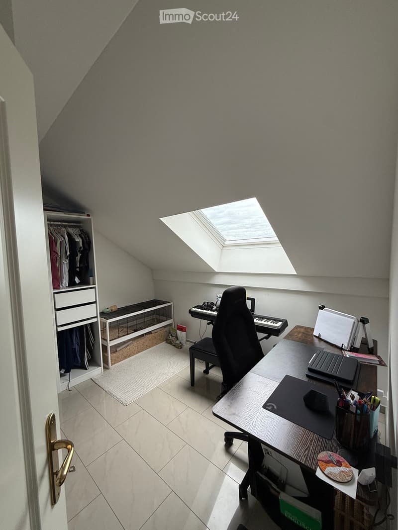 A small, well-lit room with a slanted ceiling and skylight, featuring a desk with a laptop, keyboard, mouse, and various office supplies, a chair, a piano, a pet cage, and a wardrobe with clothes.