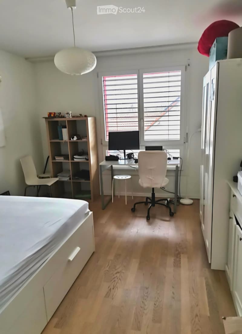 A furnished room with a bed, desk, chair, laptop, screen, shelf, and lamp. The room has wooden flooring and large windows with blinds.