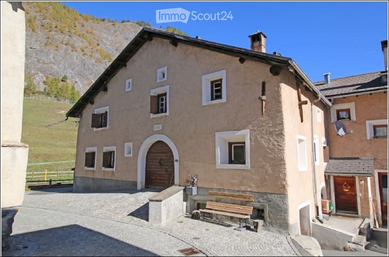 A rustic, two-story building with a beige facade, multiple windows, and a wooden door. A wooden bench is placed near the entrance, and the building is situated in a scenic, mountainous area with a clear blue sky.