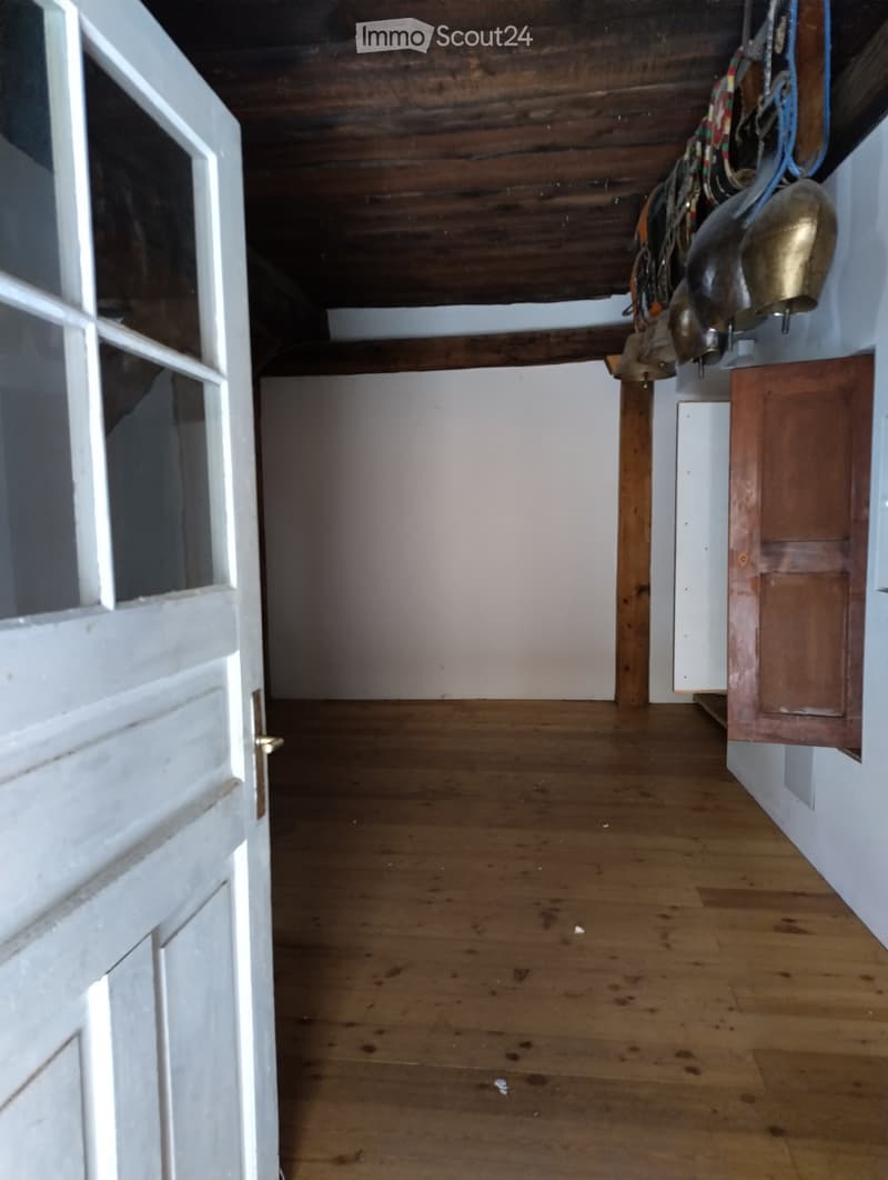 Empty wooden-floored hallway with white walls, exposed wooden ceiling beams, and a partially open white door on the left.