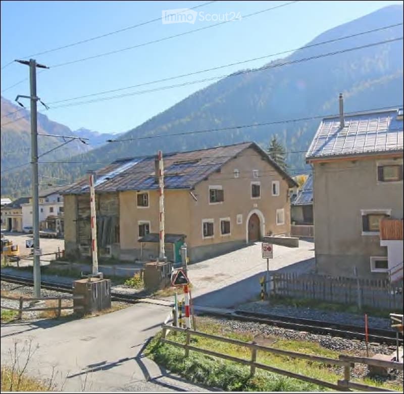 A small village scene with a two-story building featuring a tiled roof and multiple windows, surrounded by other structures and a mountainous backdrop.