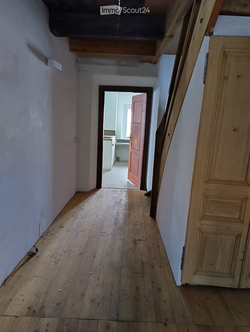 Empty wooden-floored hallway with white walls, leading to an open door revealing a kitchen area.
