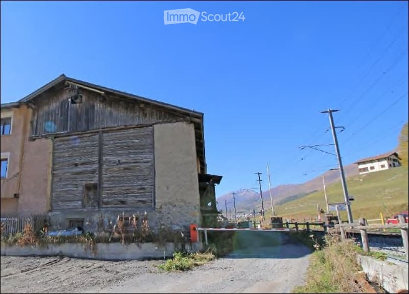 A rustic wooden and stone building with a sloped roof, situated in a rural area with mountains in the background.