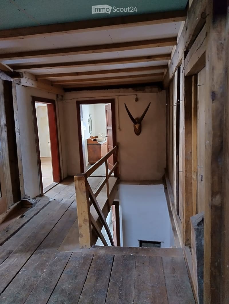 Empty wooden hallway with exposed beams, a staircase leading downwards, and a mounted animal head on the wall. The floor is made of wooden planks, and the walls are unfinished with visible wooden framing.