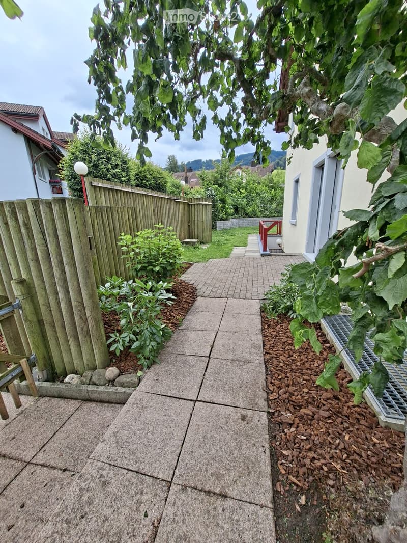 A narrow concrete pathway bordered by a wooden fence on one side and a house on the other, with small plants and mulch beds along the edges.