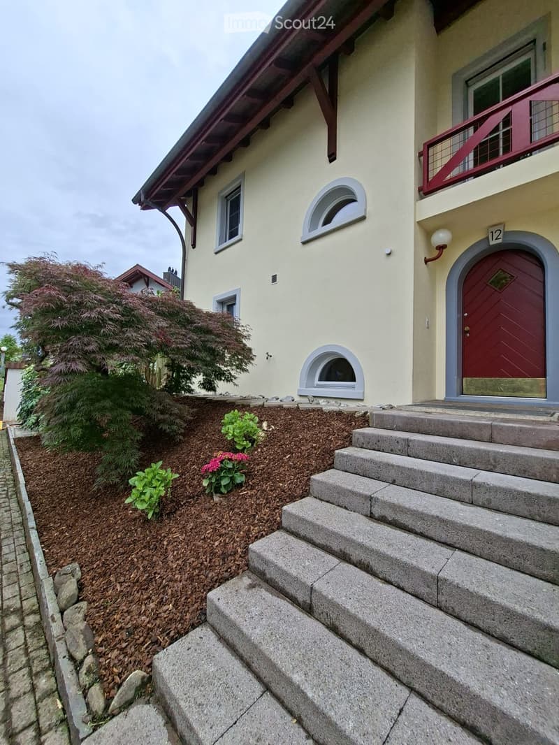 Two-story house with beige walls, red door, and small balcony with red railing. Steps lead up to the entrance, and the garden features mulch, shrubs, and flowers.
